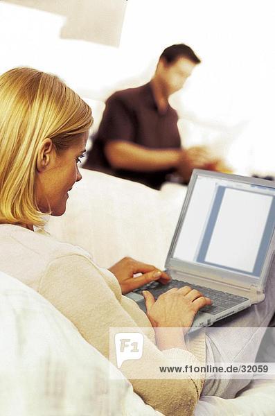 Rückansicht des Woman using Laptop auf Sofa mit Mann im Hintergrund
