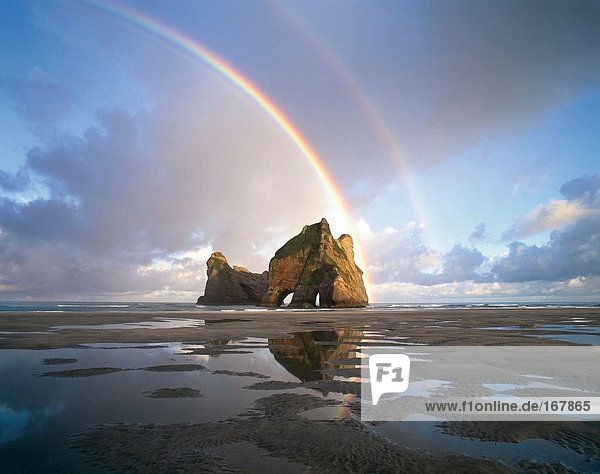Travel. New Zealand. South Island. Spit. Multiple rainbows over rock formation on beach.
