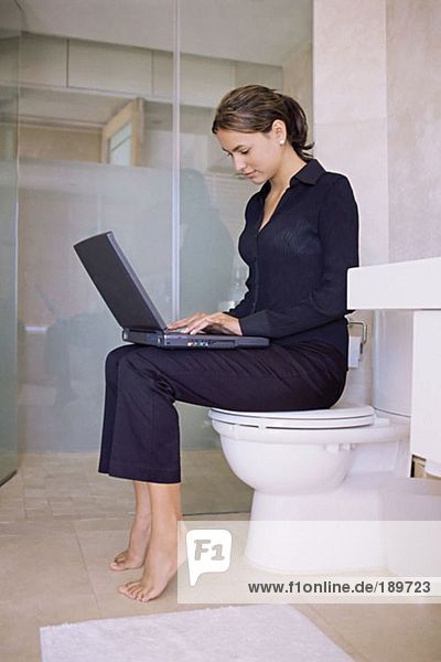 Frau sitzt auf einer Toilette und benutzt einen Laptop.