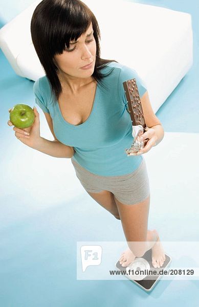Junge Frau mit Tafel Schokolade und Apfel auf Waage stehend
