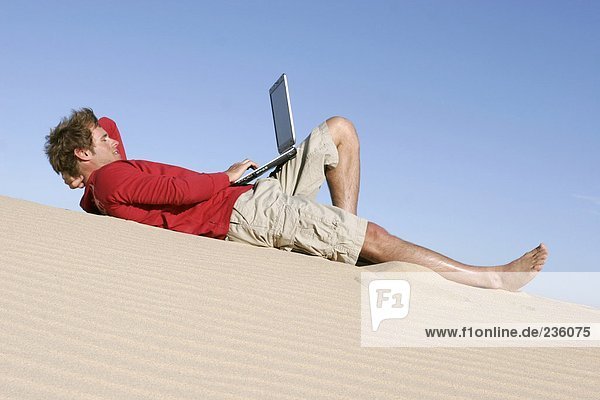 Mann auf einer Düne liegend mit einem Laptop