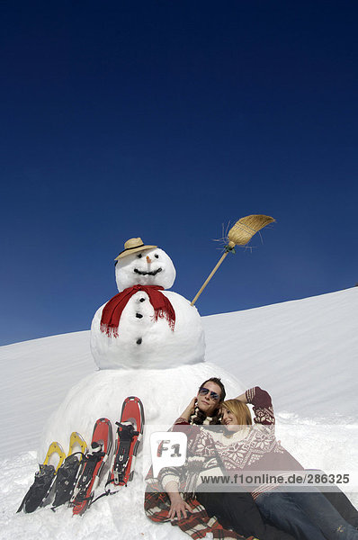 Paar sitzend bei Schneemann mit Schneeschuhen