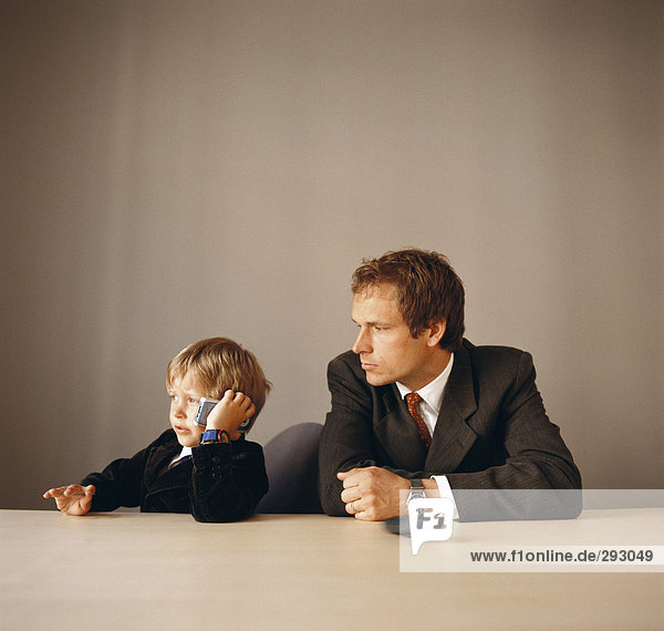 A man and a boy in suits in an office the boy talking on a mobile phone.