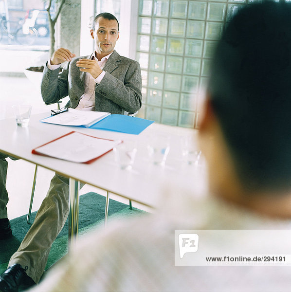 Zwei Männer sitzen im Gespräch von einer Tabelle in einer Büroumgebung.