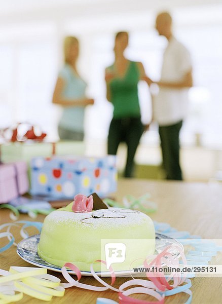 A marzipan-covered cake and presents on a table three people in the background.