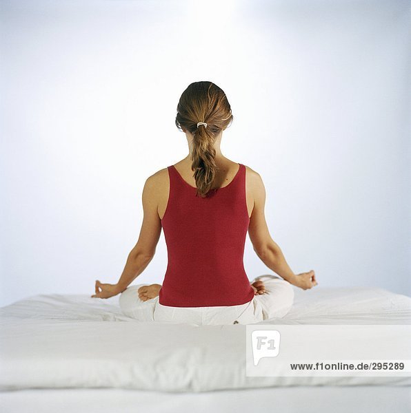 Eine Frau in Lotus Haltung auf ein Bett Rückansicht sitzend.
