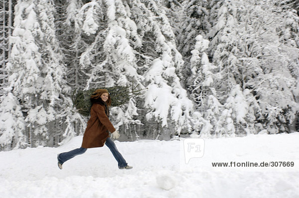 Junge Frau mit Weihnachtsbaum auf Schultern im Schnee  lächelnd