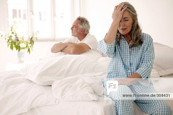 Erwachsenes Paar auf dem Bett sitzend (Fokus auf Frau im Vordergrund mit Kopf in den Händen)