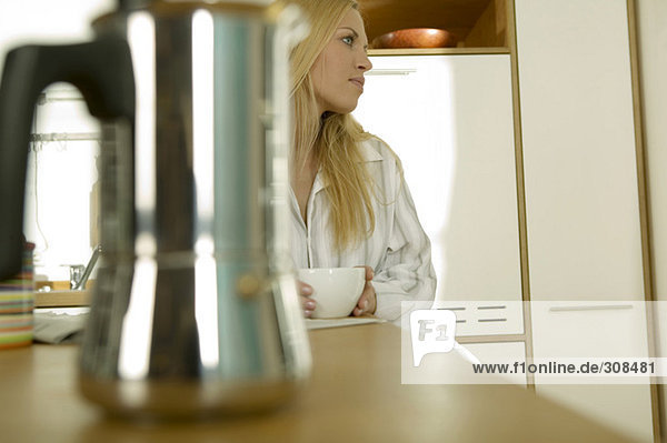 Junge Frau sitzt in der Küche mit einer Tasse Kaffee und schaut weg.