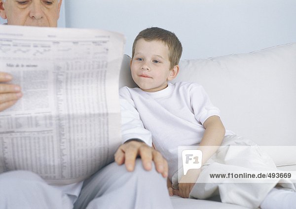 Boy sitting on sofa next to grandfather while he reads newspaper