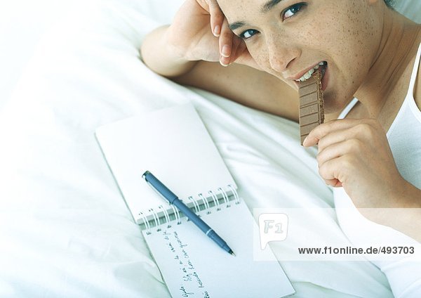 Frau liegt auf dem Bett vor offenem Notizbuch und isst Schokolade.