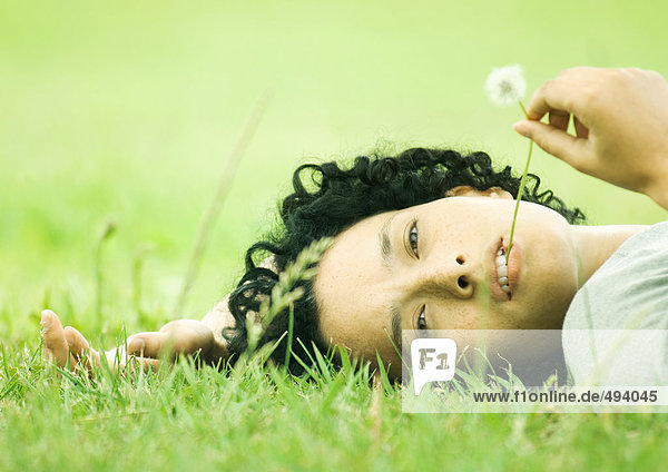 Frau auf Gras liegend mit Löwenzahn im Mund