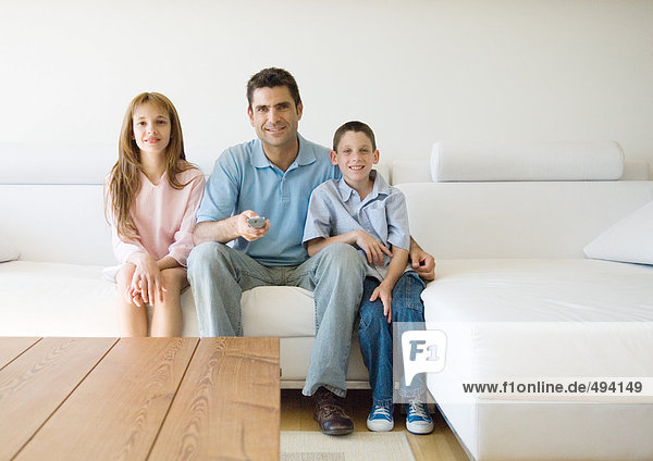 Vater und zwei Kinder sitzen auf dem Sofa und zeigen auf die Fernbedienung.