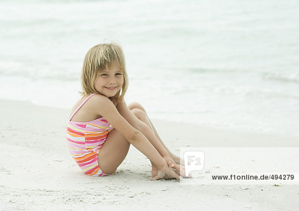 Kleines Mädchen sitzt am Strand mit erhobenen Knien und lächelt in die Kamera.