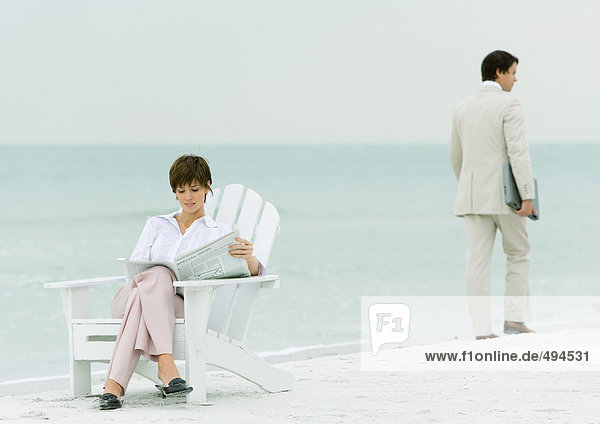 On beach  businessman carrying laptop  walking by woman reading newspaper in chair