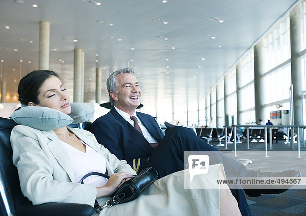 Business travelers sitting in airport lounge  woman napping with neck pillow next to smiling man