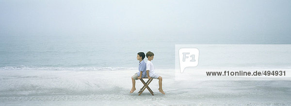 Am Strand sitzen zwei Jungen Rücken an Rücken auf einem Hocker.