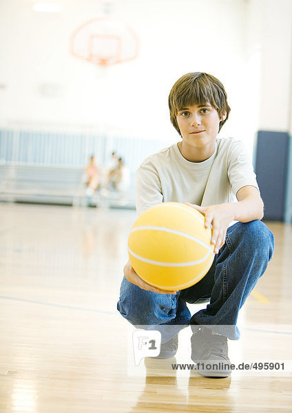 Jugendlicher Junge hockt mit Basketball in der Schulsporthalle