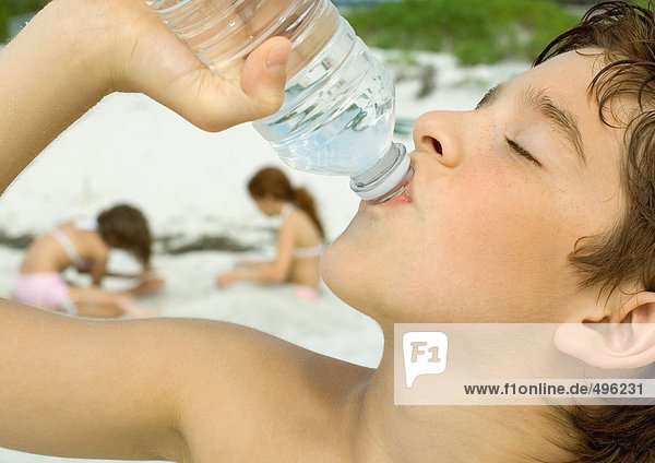 Junge trinkt Wasser in Flaschen  Mädchen spielen im Sand im Hintergrund
