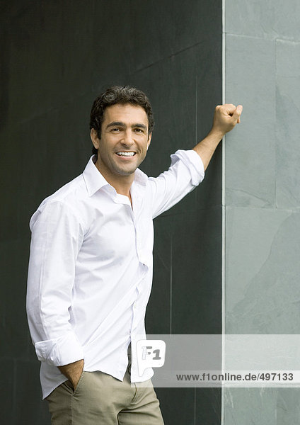 Mann steht mit der Hand an der Wandecke  lächelt in die Kamera  Porträt