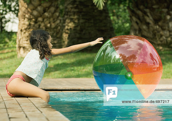 Girl sitting on edge of pool  reaching for beach ball in water