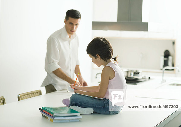 Child sitting on kitchen counter  doing homework  father in background