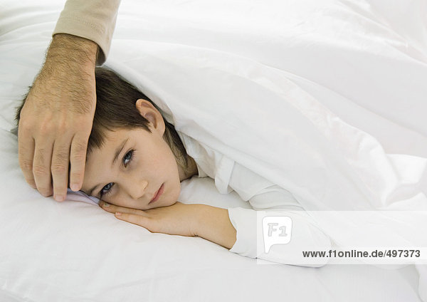 Kind im Bett liegend mit Vaters Hand auf der Stirn