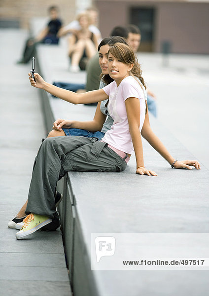Zwei Teenager-Mädchen sitzen auf einer Bank in der Stadt und fotografieren mit dem Handy.