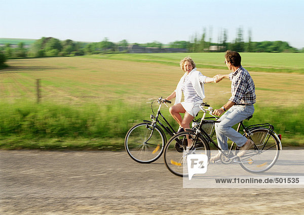Man and woman riding bicycles  holding hands  countryside in background