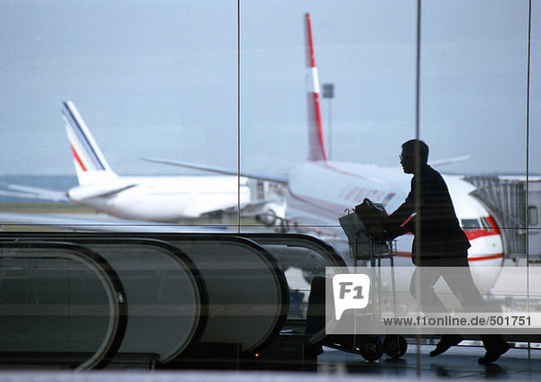 Silhouette des Mannes,  der den Gepäckwagen schiebt,  Blick durch Glasfenster,  Flugzeuge im Hintergrund