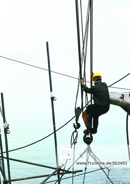 Mann mit Schutzhelm  der sich an Kabeln über Wasser festhält.