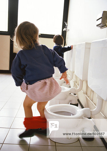 Kleines Mädchen neben der Kindertoilette mit Höschen um die Knöchel  Rückansicht  andere Toiletten und Kind im Hintergrund.