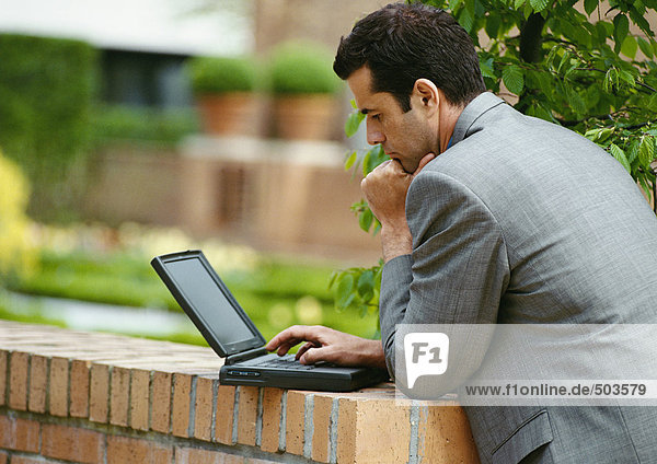 Geschäftsmann mit Laptop im Freien  lehnt sich an die niedrige Ziegelwand  Seitenansicht