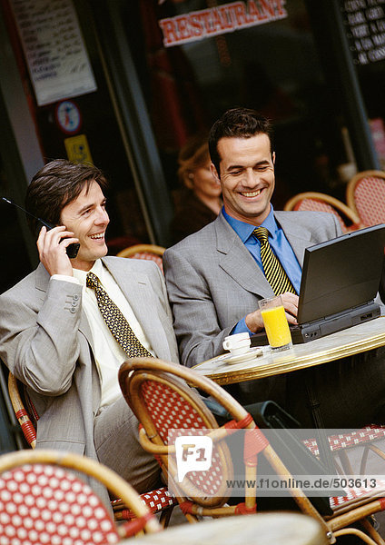 Zwei Geschäftsleute  die auf der Terrasse des Cafés sitzen und Handy und Laptop benutzen.