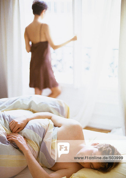 Mann im Bett liegend,  Frau am Fenster stehend im Hintergrund