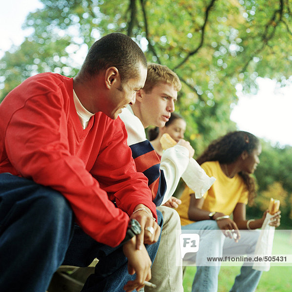 Young people sitting together on bench outside  some with sandwiches