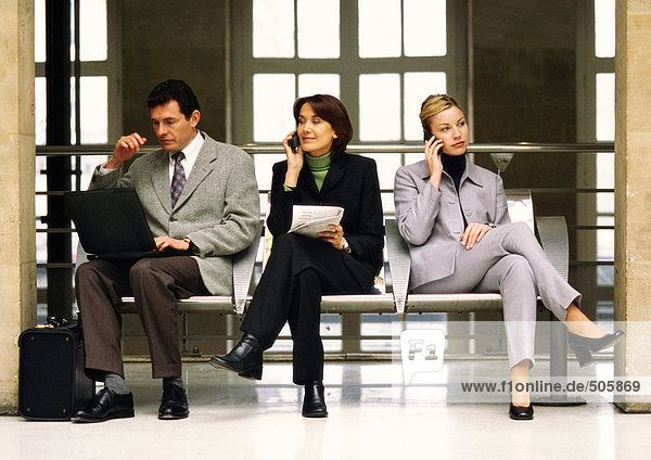 Businesswomen using cellular phones sitting next to businessman with laptop computer.