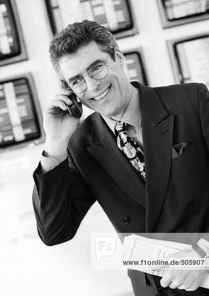 Geschäftsmann mit Handy im Flughafen  lächelnd und Zeitung vor Monitoren haltend  schwarz-weißes Porträt.