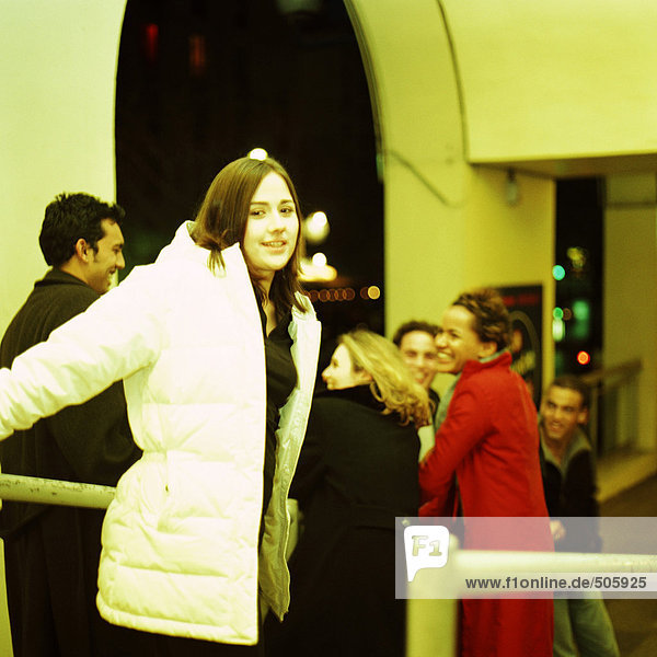 Junge Frau im Mantel lächelnd vor der Kamera,  Gruppe junger Leute im Hintergrund