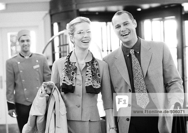Businessman and businesswoman in hotel in front of bellhop  smiling at camera  b&w.