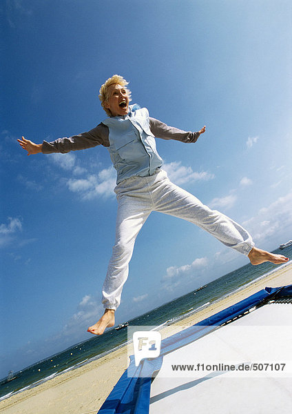 Mature woman jumping on trampoline at the beach  arms out  legs spread