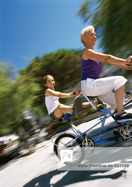 Mature woman and girl riding tandem bike  granddaugher sticking her legs out  blurred