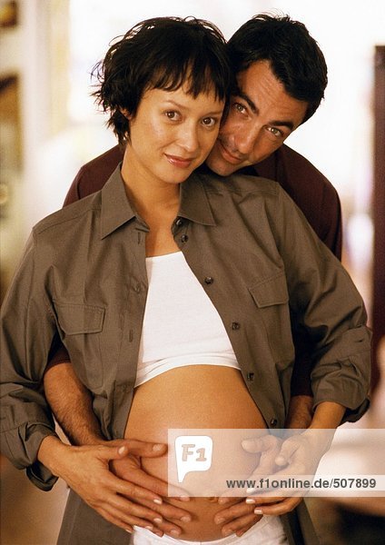 Mann berührt den Bauch einer schwangeren Frau von hinten  schaut in die Kamera  Porträt