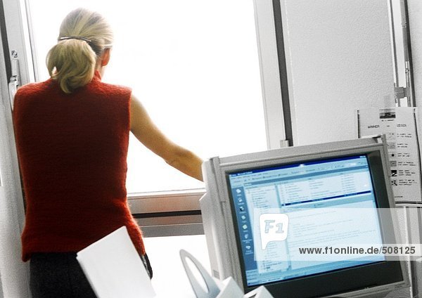 Frau im Büro stehend und aus dem Fenster schauend  Computermonitor im Vordergrund
