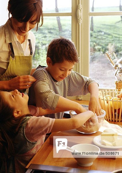 Zwei Kinder und Mutter kochen in der Küche  Kinder mit Händen in der Schüssel  Mutter in der Schürze lächelnd