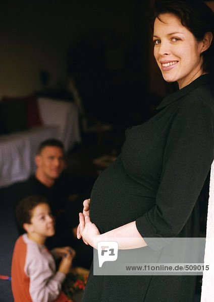 Schwangere Frau stehend und haltend  Mann und Kind sitzend im Hintergrund