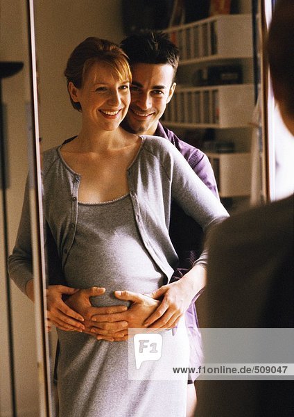 Mann mit Armen um schwangere Frau vor dem Spiegel stehend