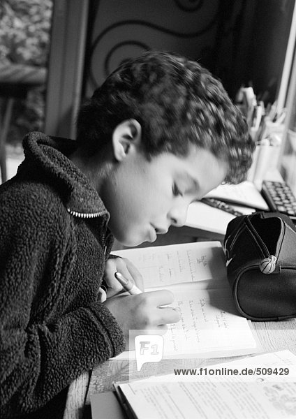 Junge sitzt am Schreibtisch  schreibt in Notizbuch  Seitenansicht  s/w