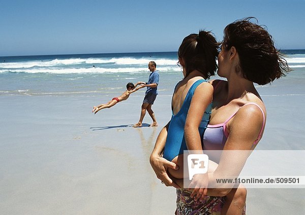 Woman holding little girl in arms in foreground on beach  man swinging little boy in background