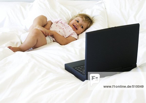 Baby auf dem Bett liegend  mit Blick auf Laptop-Computer